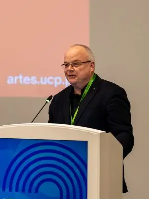 A man in a black jacket and green lanyard speaks at a podium with a HERITAGE FUTURES sign. A microphone is attached to the podium, and a screen with partial text is visible in the background.