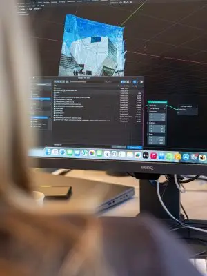 A person works on a desktop computer displaying 3D modeling software, with a building model on the screen. The viewer sees the monitor and part of the persons blurred silhouette in the foreground.