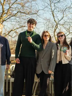 Five people stand outdoors in sunlight, smiling at the camera and holding name badges. They wear casual and business-casual clothing, with lanyards around their necks. Leafless trees and clear skies are visible in the background.