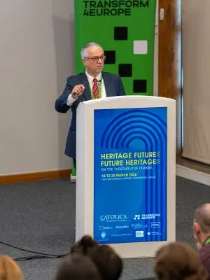 A man in a suit and tie speaks at a podium during a conference. The podium displays a blue sign for Heritage Futures: Future Heritage event. A green TRANSFORM 4EUROPE banner stands behind him. Audience members are visible.