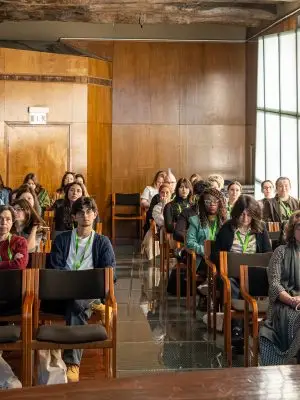 A diverse group of people sit in wooden chairs, attentively facing a speaker in a sunlit room with wood-paneled walls and large windows. Some attendees wear conference badges and green lanyards.