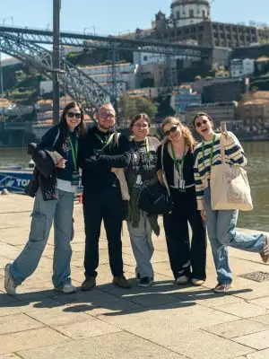 Five people stand smiling together by a river, with a bridge and historic buildings in the background. They are wearing casual clothes and lanyards, enjoying a sunny day outdoors.