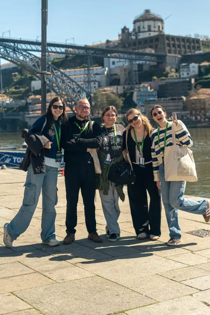 Five people smile and pose together by a river with a bridge and historic buildings in the background on a sunny day. They wear casual clothes and green lanyards, looking cheerful and relaxed.