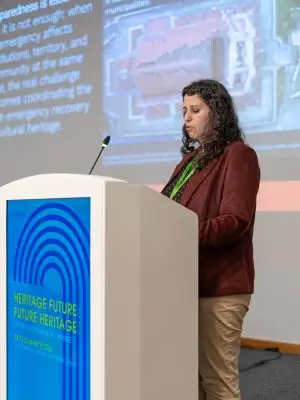A woman stands at a podium, presenting at a conference. A large screen behind her displays text and an aerial image. The podium sign reads HERITAGE FUTURES FUTURE HERITAGE 18 TO 20 MARCH 2024.