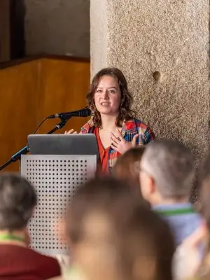A woman speaks at a podium with a microphone in front of an audience. She is wearing a red shirt and plaid jacket, and gestures with one hand as she presents. The background shows a stone wall and blurred audience members.