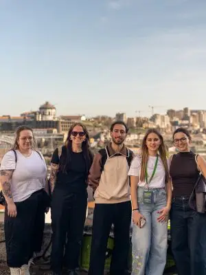 Five people stand side by side outdoors, smiling at the camera with a cityscape and blue sky in the background. They wear casual clothes, and some have tattoos and glasses.