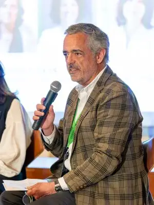 A man in a plaid blazer holds a microphone and speaks while seated at an event. He has gray hair and a beard, and is wearing a white shirt with a green lanyard. A blurred screen and other people are visible in the background.