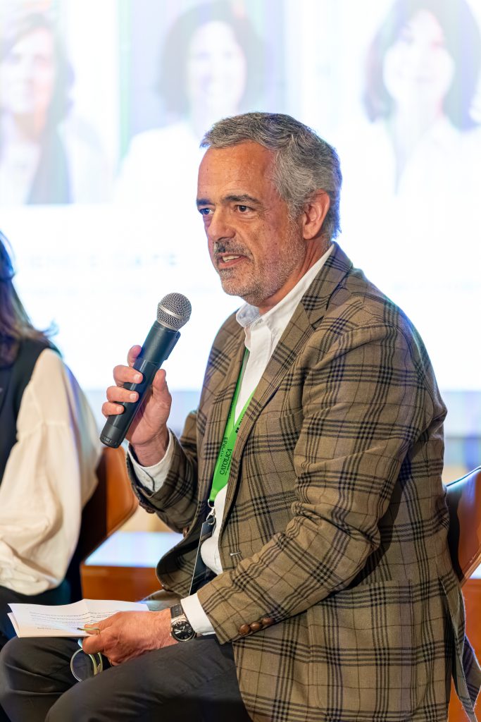 A man in a plaid blazer and white shirt holds a microphone and papers while speaking at an event. He is seated, wearing a green lanyard, with blurred people and a screen in the background.
