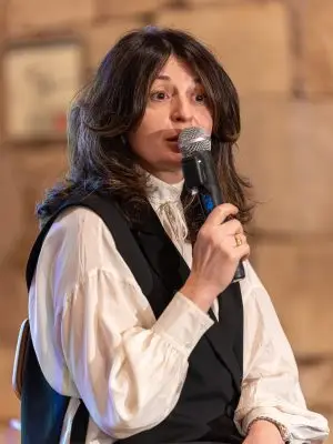 A woman with long brown hair wearing a white blouse and dark vest is holding a microphone and speaking, seated indoors with a blurred stone wall in the background.