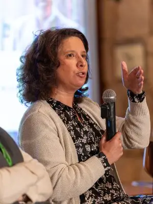 A woman with curly brown hair speaks into a microphone while gesturing with her hand. She is wearing a patterned dress and a beige cardigan, seated indoors with a blurred background.