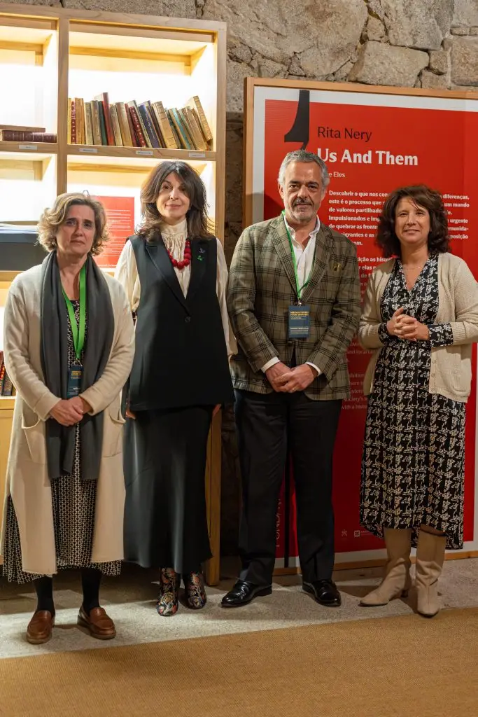 Four adults stand in front of a bookshelf and a red display panel titled Rita Nery: Us And Them. They are dressed in semi-formal attire and pose for a group photo indoors.