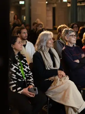 A group of people sit indoors, attentively watching something off camera. Three women in the front row are smiling; one holds a phone, another has gray hair and a scarf, and the third wears glasses and a dark outfit.