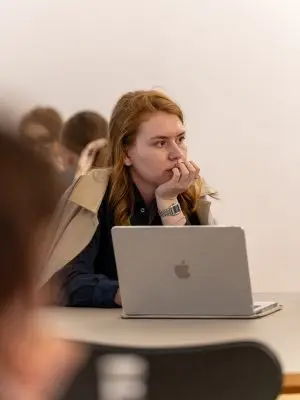 A person with long hair sits at a table, resting their chin on their hand and looking thoughtful. They are in front of an open laptop, with other people blurred in the background.