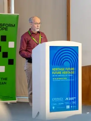 An older man with a beard and glasses speaks at a white podium with a blue sign reading Heritage Futures, Future Heritages at a conference. A green banner stands in the background.