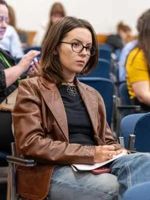 A young woman wearing glasses, a brown leather jacket, and jeans is sitting in a lecture hall, attentively listening and taking notes. Other people are seated around her, some focused, some using their phones.