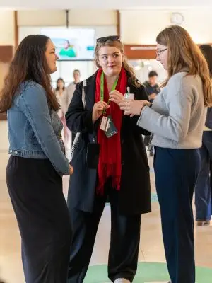 Three women stand in conversation indoors. One wears a red scarf and black coat, another a gray sweater, and the third a denim jacket. People and blurred signs are visible in the background.