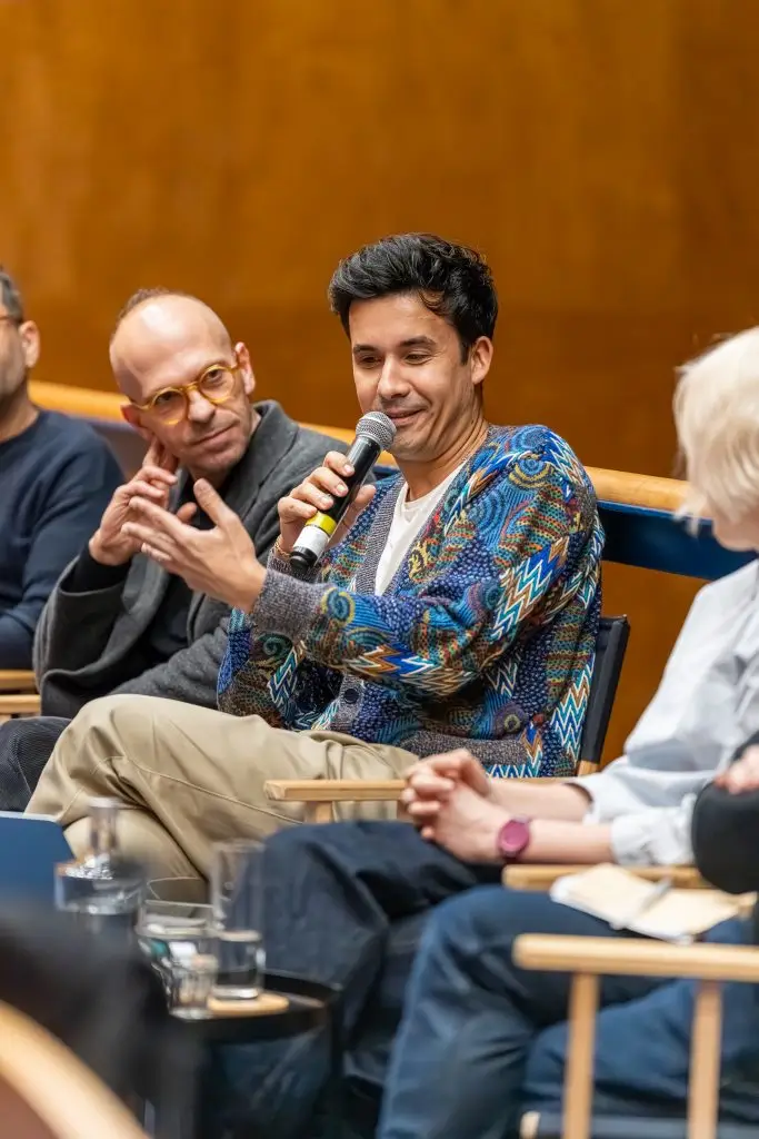 A man holds a microphone and gestures while speaking during a panel discussion with three other people seated nearby, in a well-lit indoor setting.