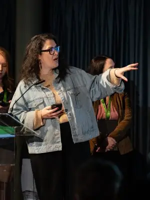 A woman wearing glasses and a denim jacket gestures to her right while speaking at a podium. Two other women stand behind her, one holding a phone and another partially visible, with dark curtains in the background.