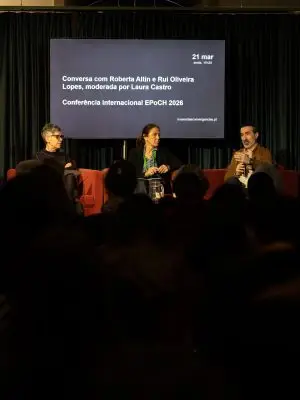 Three people sit on stage having a panel discussion, with a projected screen behind them displaying event details in Portuguese about an international conference moderated by Laura Castro, featuring Roberta Altin and Rui Oliveira Lopes.
