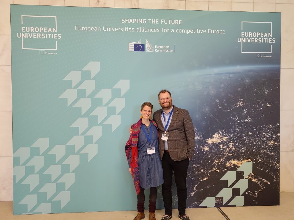 Two people standing and smiling in front of a blue backdrop with the text European Universities: Shaping the Future and graphics representing the European Union and a map of Europe.