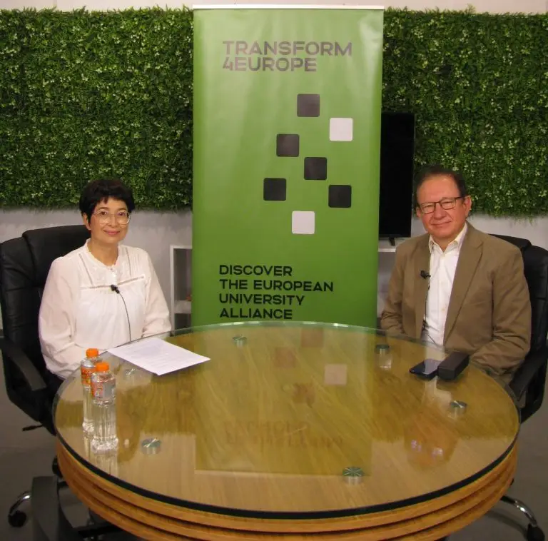 Two people sit at a round glass table with bottled water, papers, and a phone. Behind them is a green banner reading “Transform4Europe. Discover the European University Alliance,” and a wall with green foliage panels.