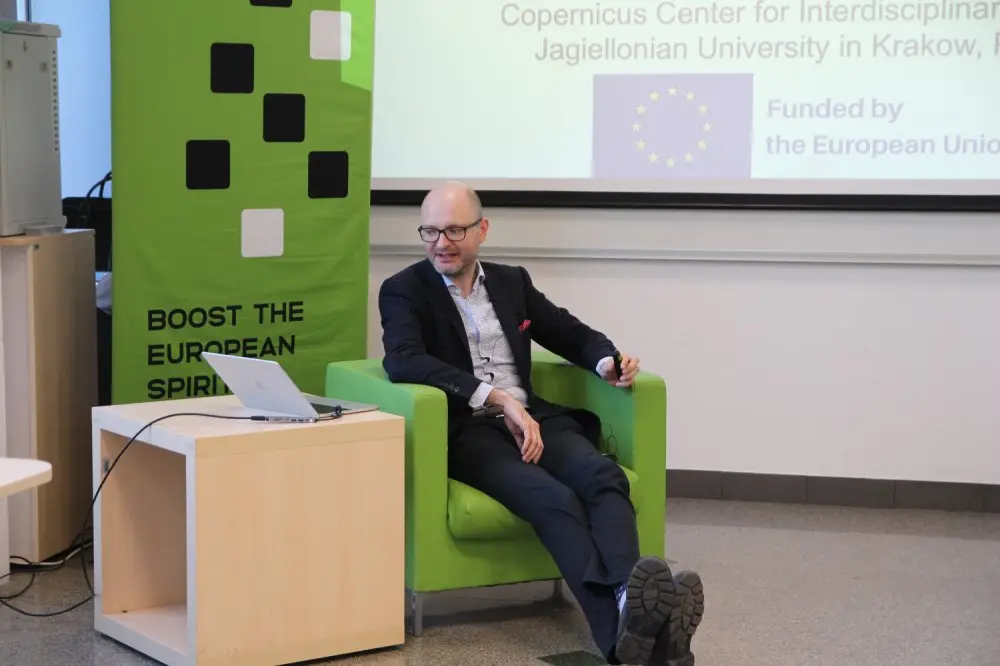 A man in a suit sits in a green chair, looking at a laptop on a small table. Behind him is a banner reading BOOСТ ТНЕ ЕUРОРEAN SPIRIT and a screen displaying funding information from the European Union.