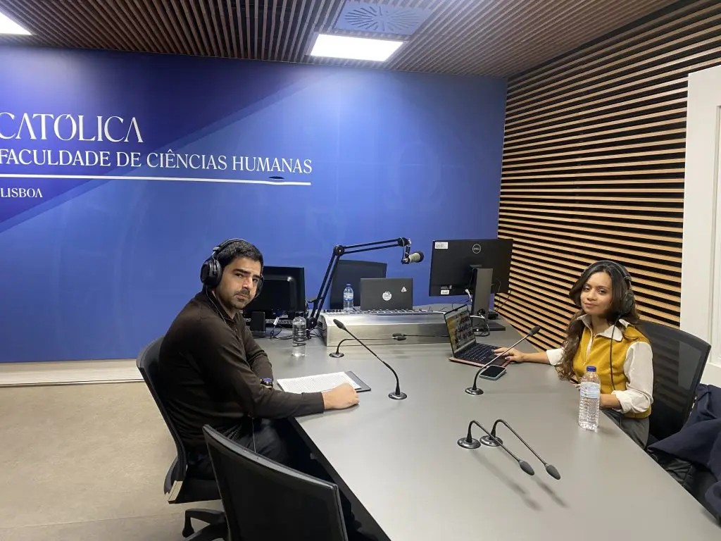 Two people wearing headphones sit across from each other at a radio studio table with microphones, laptops, and water bottles. The blue wall behind them has the text CATÓLICA FACULDADE DE CIÊNCIAS HUMANAS LISBOA.