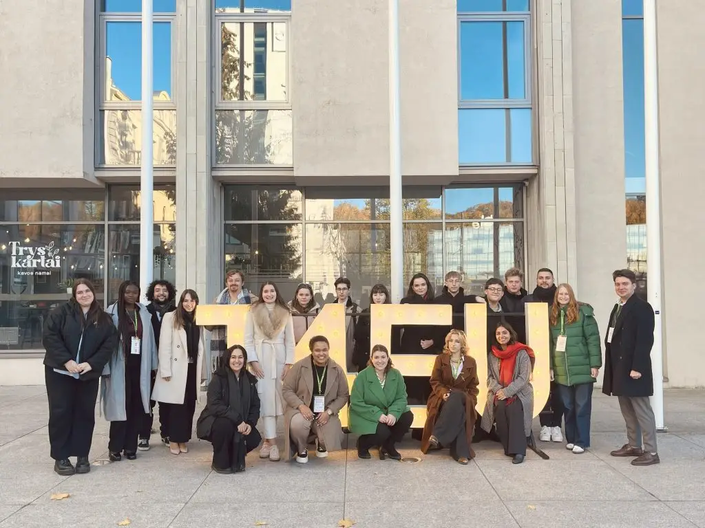 A group of twenty people poses smiling outdoors in front of a modern building with large windows. They stand and kneel around large, lit-up letters that spell ISM. Trees are reflected in the glass behind them.