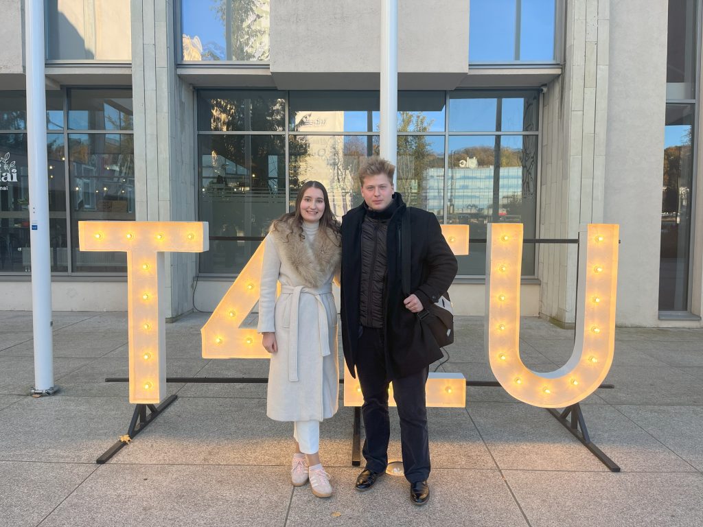 Two people stand smiling in front of large, illuminated letters that spell T4U outside a modern building with large windows on a sunny day.