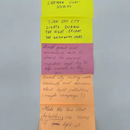 Five colorful sticky notes with handwritten text are arranged vertically on a white surface. The notes contain bullet points about city lights, animal migration, and community discussions on light pollution.