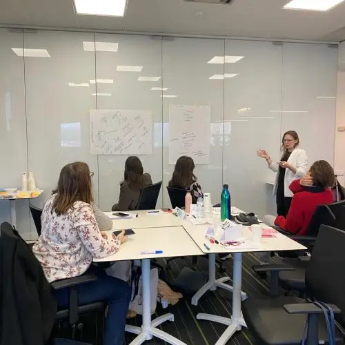 Five women are in a modern office. Four are seated around a table, while one stands and gestures toward diagrams on the glass wall, leading a presentation or discussion. Papers, cups, and markers are on the table.
