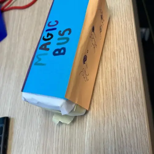 A homemade Magic Bus made from a small rectangular box, covered with blue and orange paper, with MAGIC BUS written on one side and stick figure drawings on the orange paper, placed on a wooden desk.