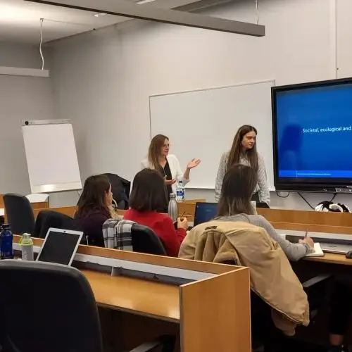Two women stand at the front of a classroom giving a presentation to seated students. A large screen displays a slide titled Società, sociologia ed…(visible text ends). Laptops and notebooks are on desks.