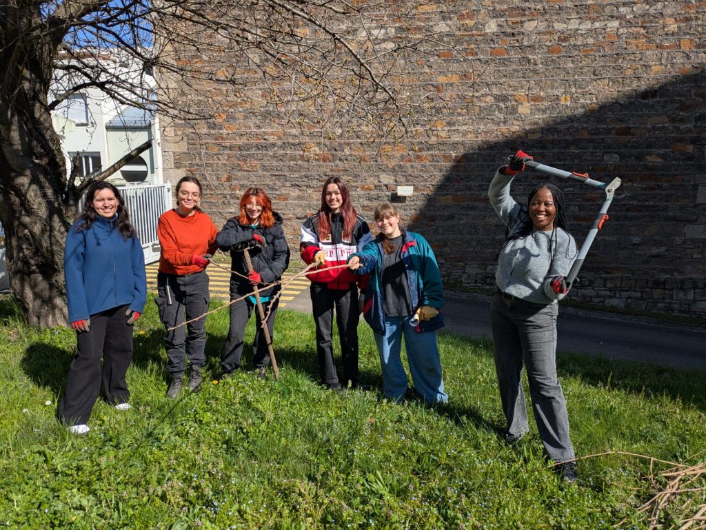 Six people stand on grass in front of a stone wall, smiling at the camera. Some are holding gardening tools like loppers and rakes, suggesting they are doing outdoor gardening or volunteer work on a sunny day.