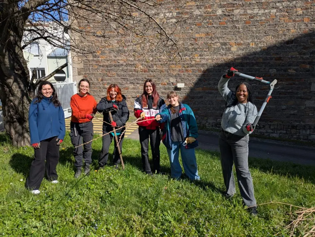 Six people stand on grass in front of a stone wall, smiling at the camera. Some are holding gardening tools like loppers and rakes, suggesting they are doing outdoor gardening or volunteer work on a sunny day.
