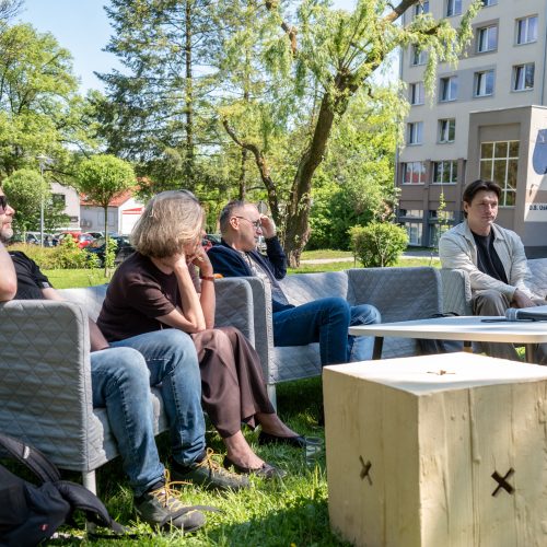 Five people sit on outdoor sofas in a grassy area beside a building, participating in a discussion. One woman speaks into a microphone while others listen attentively. Trees and parked cars are visible in the background.