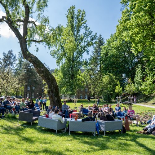 A group of people sit on sofas and chairs arranged in a circle on a grassy field under tall trees, enjoying a sunny day outdoors. Some are talking, while others relax in the shade.