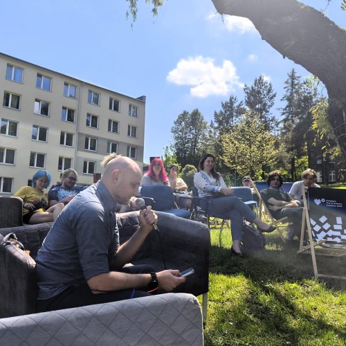 A group of people sits outdoors on chairs and couches near a large tree, some listening and some talking. An apartment building is in the background, and sunlight filters through the trees.