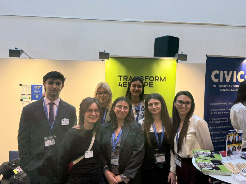 Seven young adults, dressed in business attire and wearing conference badges, stand together and smile at a booth with a bright green Transform 4Europe banner and informational displays in the background.