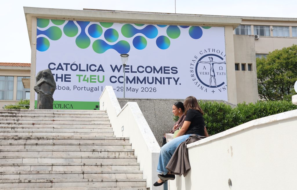 Two people sit on white outdoor stairs near a large banner that reads CATÓLICA WELCOMES THE T4EU COMMUNITY. Lisboa, Portugal, 27–29 May 2026 on a university building.