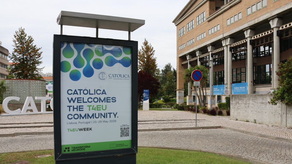 An outdoor sign reads “CATÓLICA welcomes the T4EU community. Lisboa, Portugal | 23-29 May 2026” with university buildings, trees, and pathway benches in the background.
