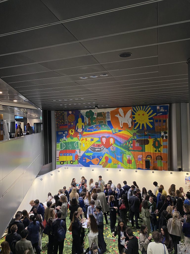 A large group of people gathers in a modern indoor space beneath a colorful mural featuring abstract animals, people, and a white dove. The ceiling is metallic and the atmosphere appears lively and social.