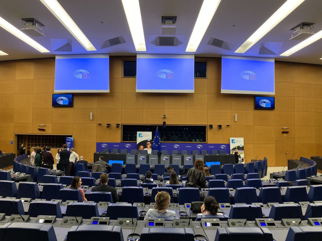 People gather in a modern, mostly empty conference room with rows of blue seats facing a stage with multiple podiums. Large screens display a blue presentation slide at the front of the room.