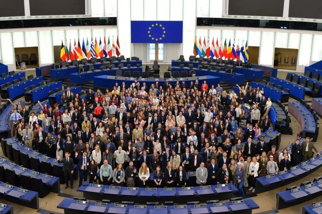 A large group of people pose together in the center of a circular parliamentary chamber with blue seats, surrounded by desks and flags of European countries, beneath a large European Union flag.