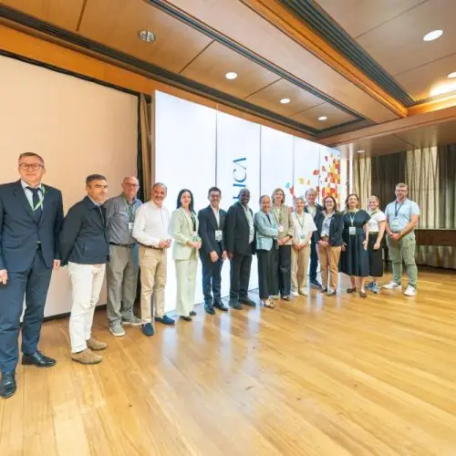 A group of people stands in a line on a wooden stage in a conference room, with a large white screen and conference banner behind them. One person stands slightly apart from the group on the left.
