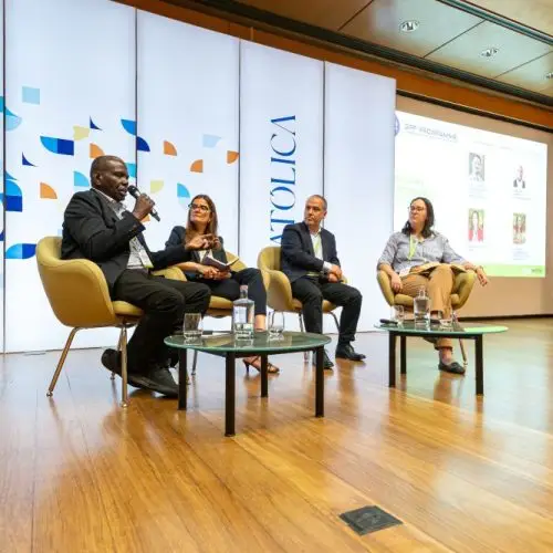 Four people sit on stage in armchairs, engaged in a panel discussion. Behind them is a large display with patterns and the word CATOLICA. A screen to the right shows presentation slides.
