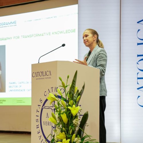 A woman stands at a podium decorated with yellow flowers, speaking at an event. Behind her, a screen displays a presentation slide titled A Choreography for Transformative Knowledge with a photo and text.