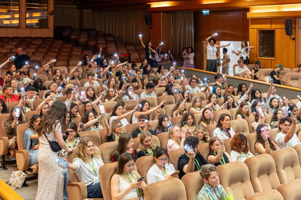 A large group of people sit in an auditorium, many holding up phones with flashlights on, creating a lively atmosphere. The audience appears engaged and cheerful, with some standing and taking photos.