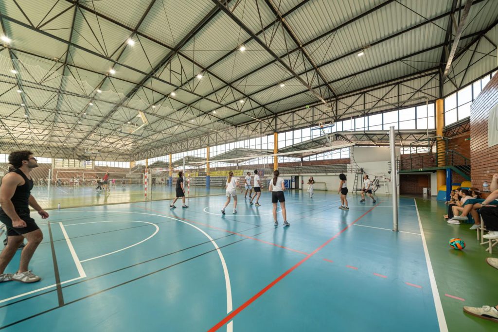 A group of people play indoor volleyball in a spacious gym with high ceilings and large windows. Some players are mid-action at the net, while others and spectators sit on the side benches.