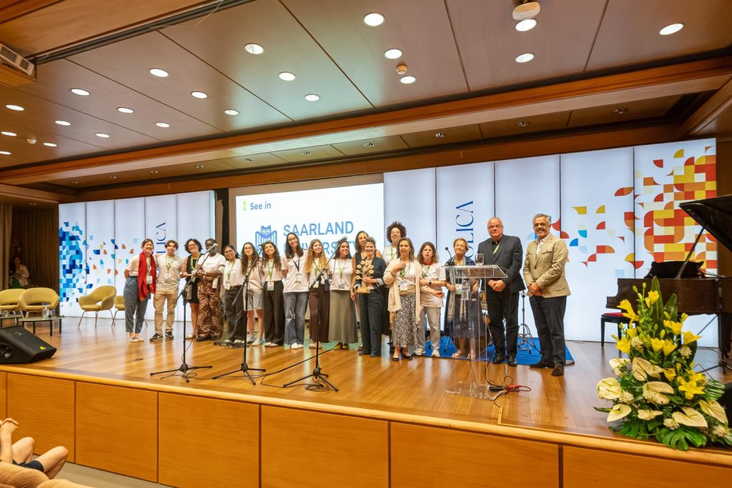 A group of people stand on a stage in front of a podium and large display screens that read SAARLAND. The setting appears to be a formal event or conference, with several people smiling for a group photo.